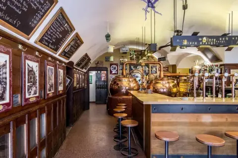 Interno di un locale tradizionale con alambicchi di rame, sgabelli e insegne che richiamano il mondo della birra e del **hopfen & co.**