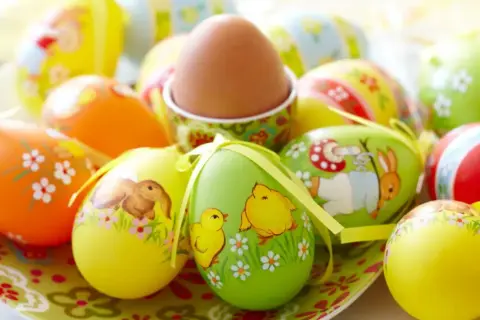 Un assortimento di colorate decorazioni uova di Pasqua con coniglietti, pulcini e fiori. Un uovo marrone è in un portauovo decorato.