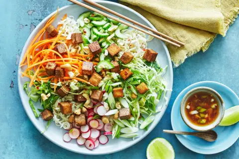 En färgglad asiatisk nudelsallad med krispiga grönsaker, cashewnötter och gyllene tofu-kuber. Serveras med en dressing och limeklyftor.