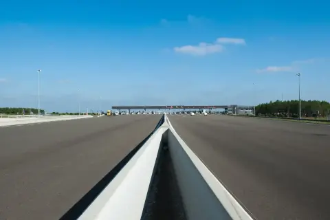 Péage autoroutier sous un ciel bleu. La route s'étend vers une barrière de péage avec plusieurs voies. Un camion jaune est visible.