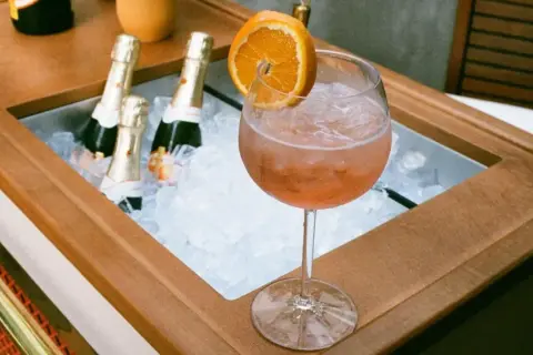 Orzeźwiający drink z prosecco i colą, ozdobiony plasterkiem pomarańczy, obok schłodzonych butelek.