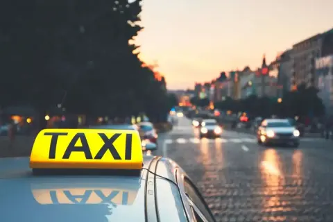 Żółty znak TAXI na dachu samochodu na tle miasta o zachodzie słońca.