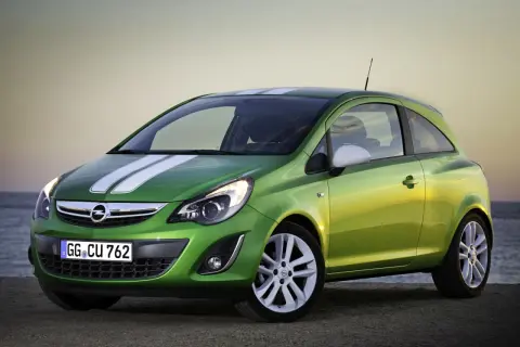 Zielony Opel Corsa D z białymi pasami na masce i dachu, zaparkowany na plaży o zachodzie słońca.
