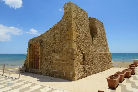 Antica torre di pietra sulla costa, con il mare azzurro sullo sfondo. Vicino, una fila di vasi di terracotta, come tre fontane in attesa.