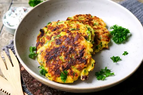 Goldbraune Zucchini-Puffer, garniert mit Petersilie, liegen auf einem Teller. Daneben eine Schüssel mit Dip.
