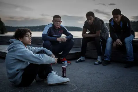 Fyra unga män sitter vid vattnet i skymningen, en scen från filmen Vinterviken (2021).