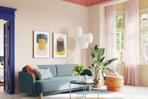 Ein Wohnzimmer mit blauem Sofa, Kunstwerken und einem orangefarbenen Hocker. Die Decke ist in einem kräftigen Pink gestrichen, was einen schönen Kontrast zur hellen Wandfarbe bildet.
