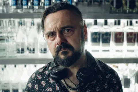 Dr Misio, znikam. Mężczyzna z zarostem i słuchawkami na szyi stoi przed lodówką pełną butelek.