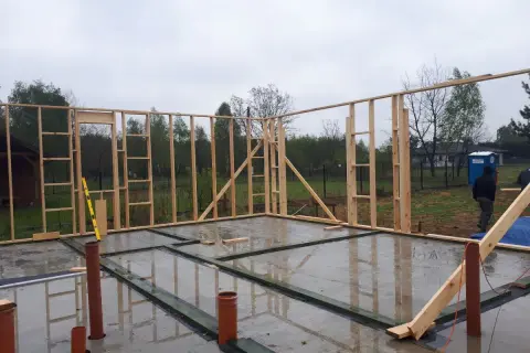 Budowa fundamentu pod dom szkieletowy. Drewniana konstrukcja ścian stoi na betonowej płycie, widać rury kanalizacyjne.