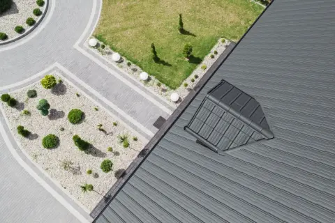 Nowoczesny dach pulpitowy z szarej blachy, z widocznym oknem dachowym. Obok zadbany ogród z brukowanymi ścieżkami i niską roślinnością.