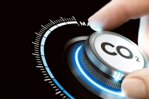 Dłoń obraca pokrętło z napisem CO2, zmniejszając emisje. Skala wskazuje na redukcję.