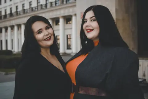 Dwie uśmiechnięte kobiety, uosobienie body positivity, pozują przed budynkiem z kolumnami.