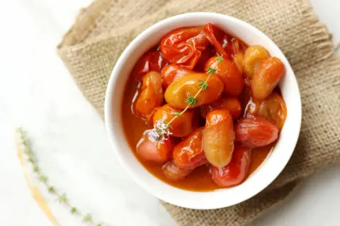 Deliciosos tomates cherry confitados en un cuenco blanco, adornados con tomillo fresco y una flor morada.