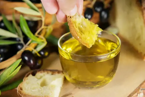 Un pezzo di pane intinto in olio dorato, con olive e rami d'ulivo sullo sfondo, evoca le antiche origini olio di oliva.