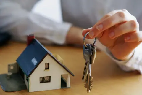 Mano sostiene llaves frente a maqueta de casa. Una posible solución si un heredero no quiere vender.