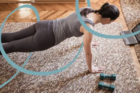 Kobieta w pozycji deski ćwiczy pilates w domu, patrząc na laptopa. Obok leżą hantle.
