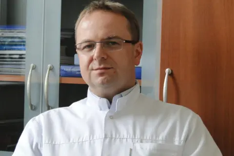 Portret mężczyzny w okularach i białym fartuchu lekarskim, na tle regałów z książkami.
