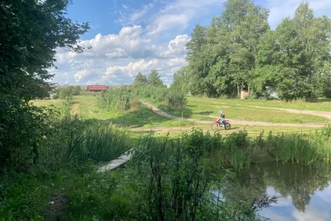 Osoba na motocyklu crossowym jedzie po szutrowej drodze wśród zieleni, obok stawu i drewnianego mostku.