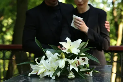 Białe lilie na ciemnym blacie, w tle para w żałobie.