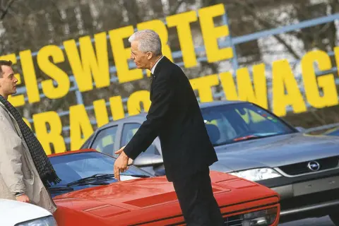 Dwóch mężczyzn ogląda sportowe auta. Jeden wskazuje na czerwony samochód, drugi stoi obok.