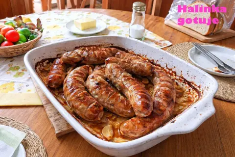 Pyszna, pieczona kiełbasa biała w naczyniu żaroodpornym, z cebulką i czosnkiem. Obok koszyk z jajkami i kromka chleba.