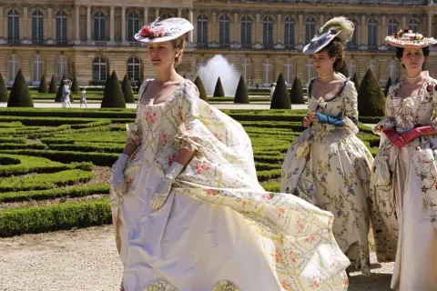 Kvinnor i magnifika **rococo fashion**-klänningar med blomstermönster promenerar i en formell trädgård framför ett palats.