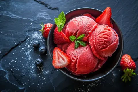 Schüssel mit erfrischendem Erdbeer-Sorbet-Eis, garniert mit frischen Erdbeeren und Minze, daneben Blaubeeren.