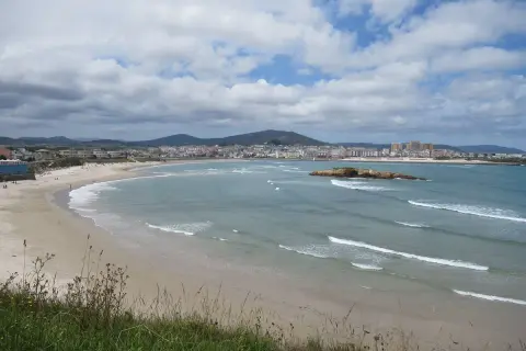 Amplia vista de la playa de Altar Barreiros con arena dorada, mar azul y una ciudad costera al fondo.