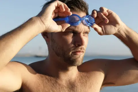 Mężczyzna zakłada niebieskie okulary pływackie na tle błękitnego nieba i wody.