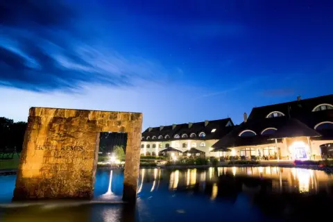 Hotel Remes Sport & Spa nocą, z kamiennym łukiem i stawem w tle.