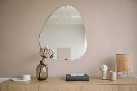 Un miroir aux formes organiques reflète la pièce. Sur le meuble en bois, une fleur séchée, des livres, une lampe en rotin et des objets décoratifs créent une scène apaisante.