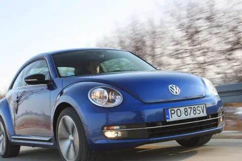 Niebieski, nowy Beetle pędzi po drodze, za nim rozmyty krajobraz drzew.