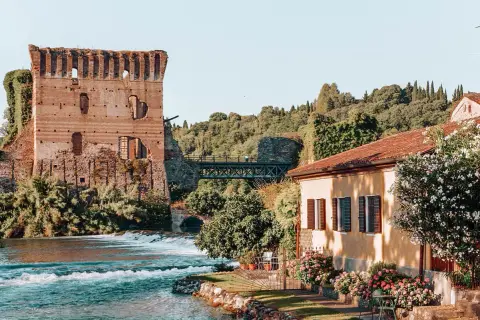Rovine di un castello medievale e una casa con fiori colorati, un fiume e un ponte: un quadro perfetto dei borghi veneti da visitare.