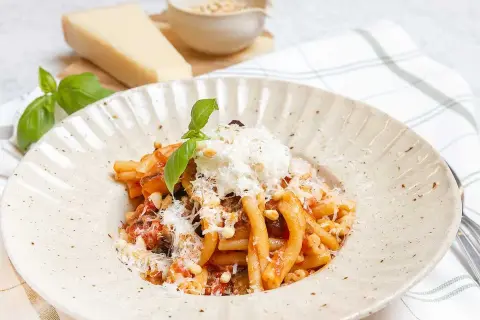 En tallrik med pasta alla norma, toppad med ricotta och basilika. Bredvid syns en bit parmesanost och en skål med pinjenötter.