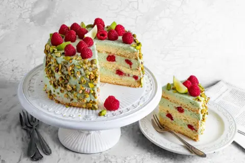 Wyjątkowy tort pistacjowo malinowy, udekorowany świeżymi malinami i pistacjami. Kawałek na talerzyku kusi warstwami kremu i owoców.