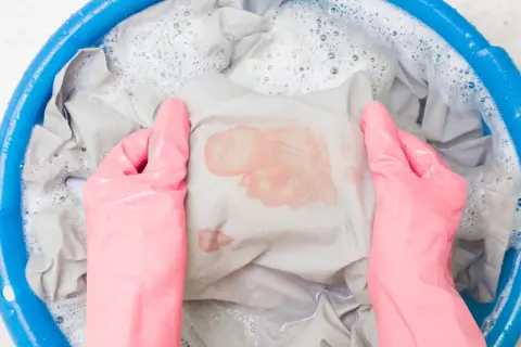 Mani con guanti rosa che lavano un vestito macchiato di rosso in una bacinella blu con acqua saponata. Ecco come sbiancare i vestiti che hanno preso colore.