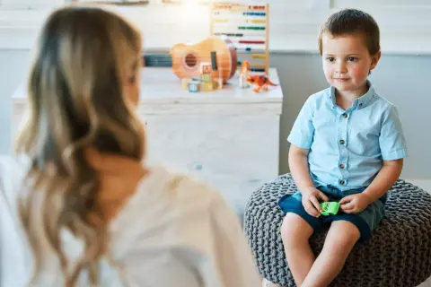 Chłopiec w niebieskiej koszuli siedzi na pufie, rozmawiając z psychologiem. W tle zabawki.
