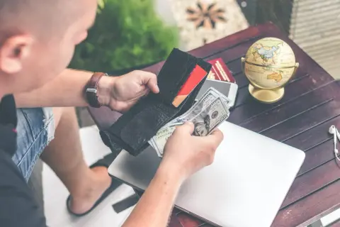 Mężczyzna liczy pieniądze w portfelu, przygotowując się do podróży. Na stole leży globus, paszport i laptop, sugerując potrzebę finansowego assistance.