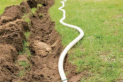 Ein Graben wird für das Erdkabel verlegen vorbereitet. Ein weißes Kabel schlängelt sich über das Gras neben dem aufgeworfenen Erdreich.