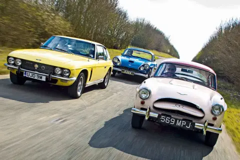 Drei klassische Autos, ein gelber Jensen Interceptor, ein blauer und ein rosafarbener, fahren auf einer Landstraße.