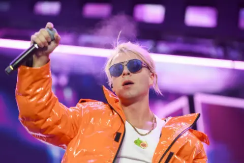 Artysta w pomarańczowej kurtce z mikrofonem, w okularach przeciwsłonecznych. Ciekawe, ile skolim bierze za koncert, że stać go na taki strój!