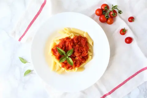 Penne pomodoro z bazylią na białym talerzu, obok gałązka pomidorków koktajlowych.