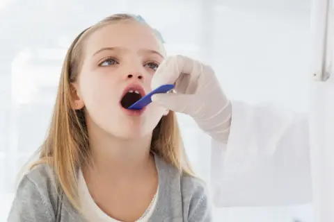 Niña con la boca abierta, examinada por un médico. Podría ser una consulta previa a una **operación amígdalas niños**.