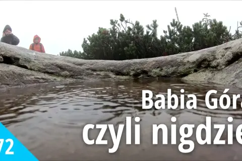 Mglisty dzień na Babiej Górze. Dwie osoby w kurtkach przeciwdeszczowych stoją za kamieniem, obok kałuży.