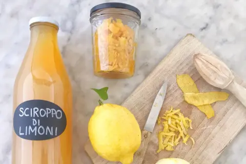 Bottiglia di sciroppo di limoni, limoni freschi, scorze e un coltello su tagliere. La sciroppo di limone ricetta della nonna è pronta!