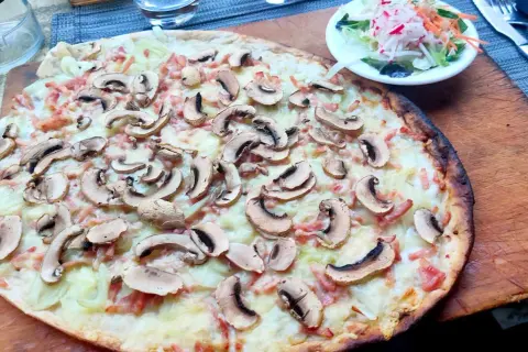 Ein köstlicher Straßburger Flammkuchen mit Pilzen und Speck, serviert auf einem Holzbrett, daneben ein kleiner Salat.