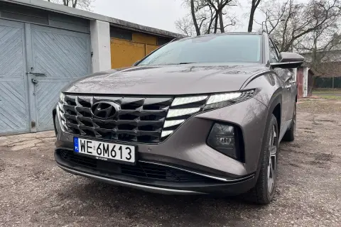 Szary Hyundai Tucson Hybrid z charakterystycznym grillem i światłami LED stoi przed garażami.