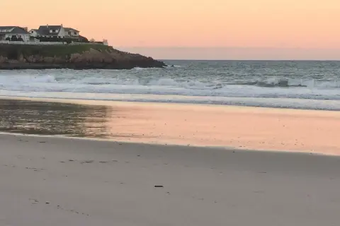Ocaso en la playa areoura. Olas rompen suavemente en la arena dorada, reflejando el cielo rosado. Casas blancas coronan el acantilado.