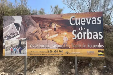 Cartel anunciando las Cuevas de Sorbas, con imágenes de exploradores y formaciones rocosas. Ofrecen visitas guiadas, restaurante y tienda.