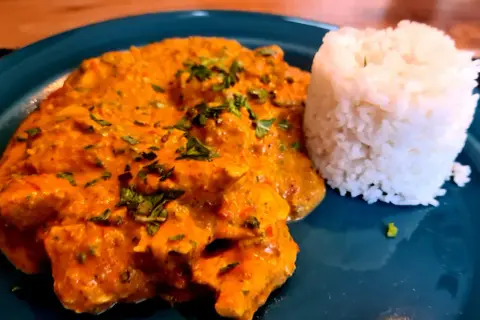 Ein Teller mit cremigem Hähnchen Tikka Masala, garniert mit frischer Petersilie, daneben ein Berg aus duftendem Reis.